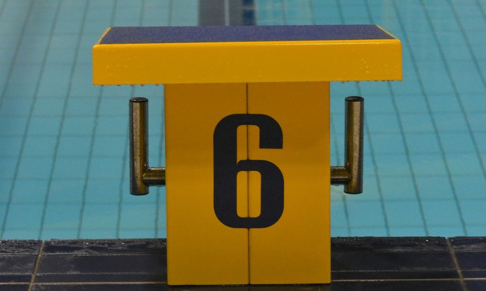 Das Foto zeigt im Vordergrung einen gelben Schwimm-Startblock mit der Nummer 6 in schwarzer Schrift. Im Hintergrund ist die Schwimmbadbahn zu erkennen. Das Bild dient als Beitragsoto für einen Blogartikel zu Starts und Wenden im freizeitsportlichen Schwimmen.