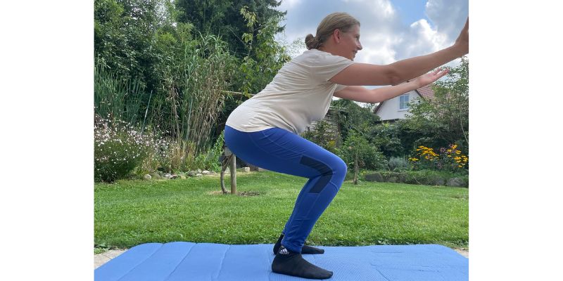 Im Vordergrund des Bildes steht eine weibliche Sportlerin auf einer blauen Iso-Matte in der für Squats üblichen Hockposition mit nach vorn ausgestreckten Armen. Die Sportlerin trägt eine blaue lange Leggins und ein kurzärmliges braun-graues T-Shirt. Im Hintergrund ist ein Ausschnitt eines Sommer-Gartens zu sehen.