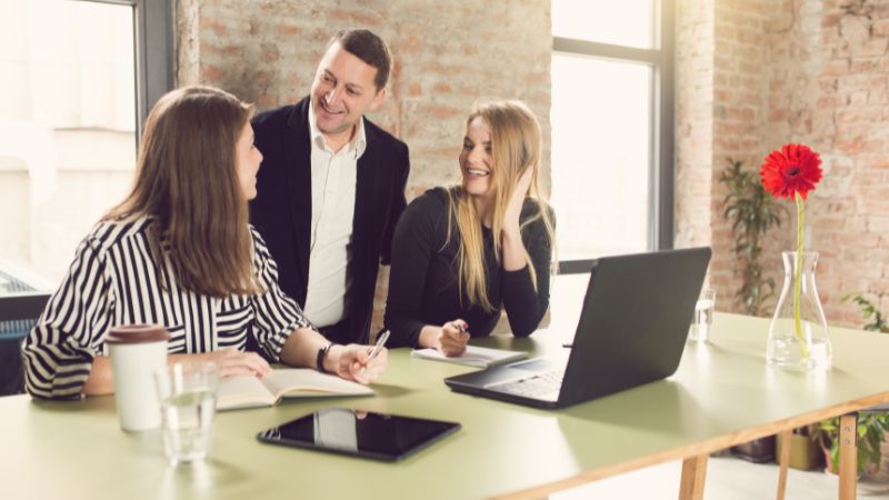 zwei Frauen und ein Mann stehen hinter einem Laptop im Büro und lächeln entspannt